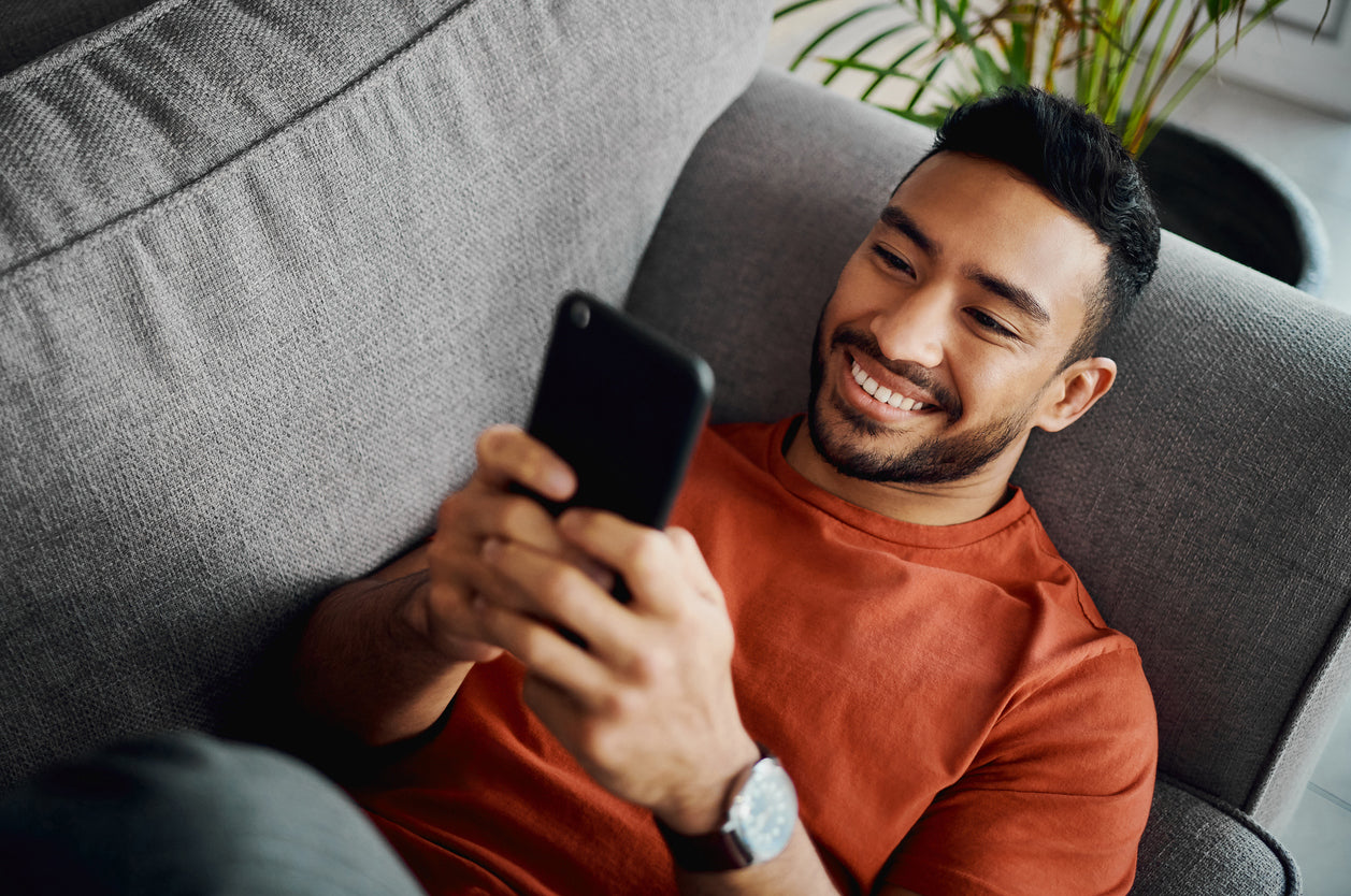 A person in a rust-colored t-shirt relaxes on a gray couch, holding a smartphone and surrounded by indoor plants.