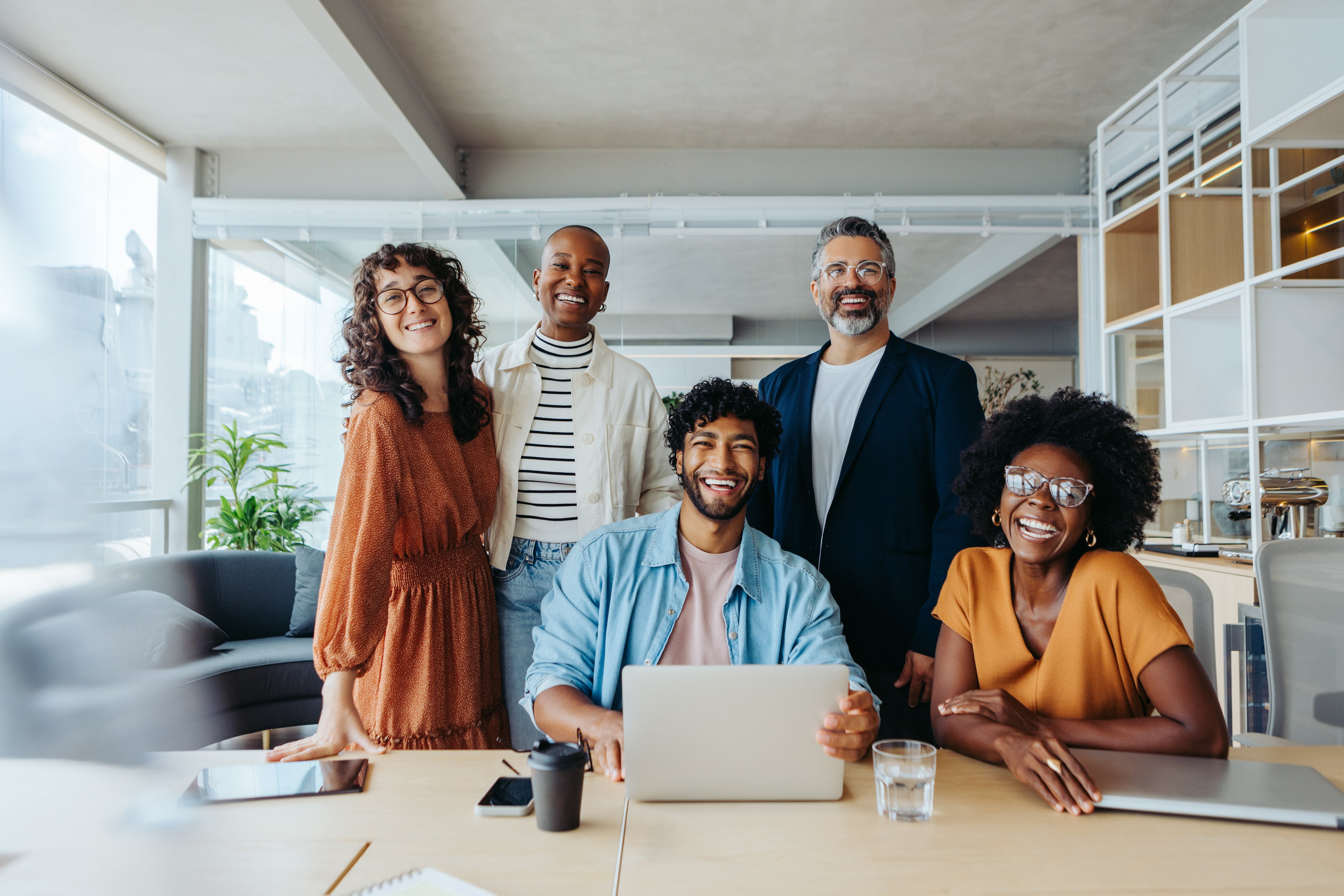 A diverse group of professionals stands around a table, with a laptop and drinks, in a bright, modern office space.